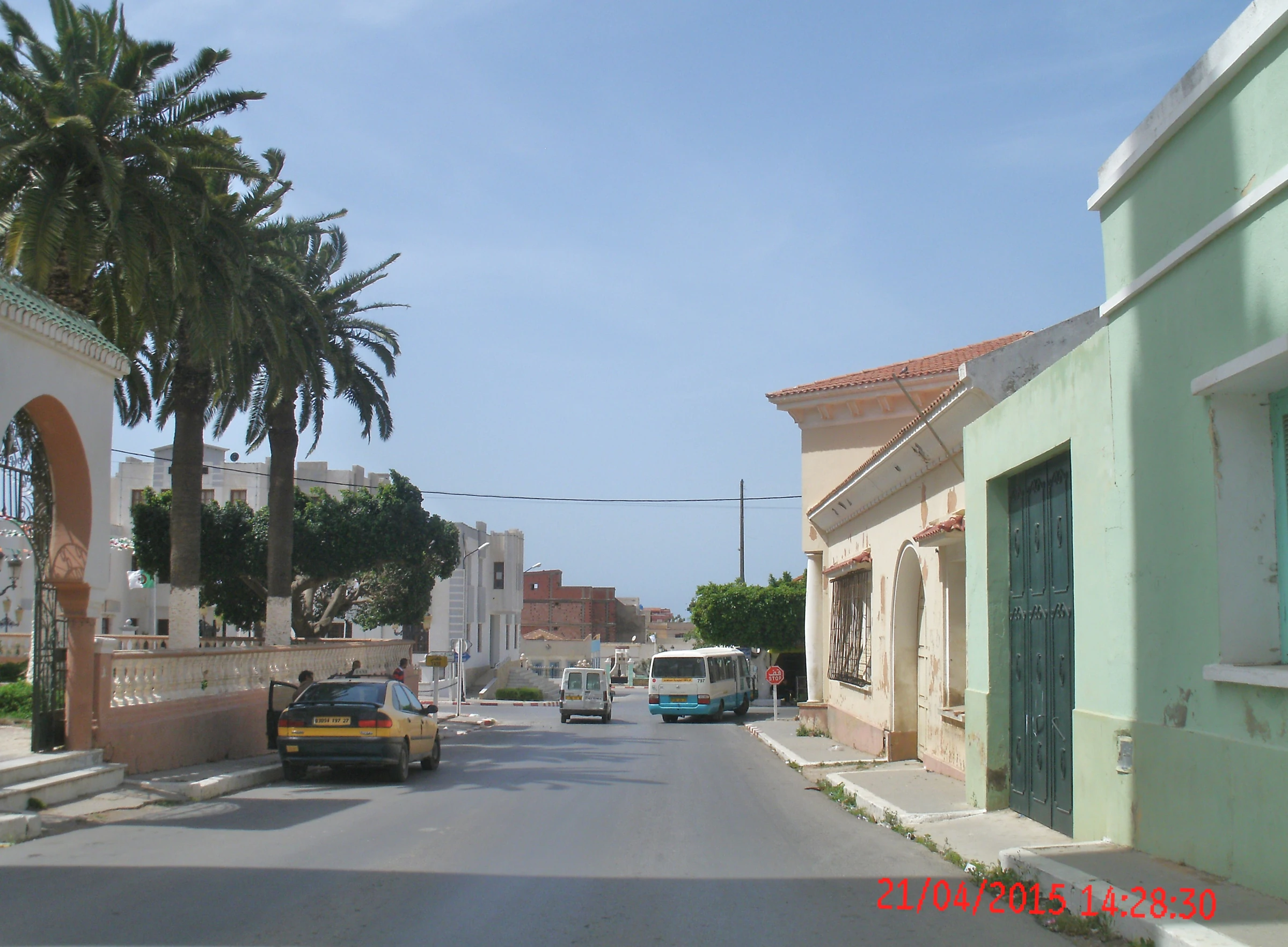 Rue de Stidia photographiée en avril 2015 : façades claires, porte verte, palmiers et circulation sur une rue en pente. Photographie documentaire de Yelles ; aucune origine architecturale allemande déduite de cette vue.