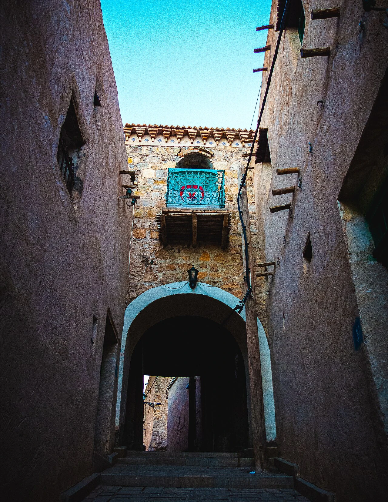 Photographie réelle d’une ruelle du ksar de Bou Saâda : murs de terre, passage voûté, petites poutres et balcon à grille turquoise portant un motif rouge, tels que photographiés par BessenouciIslem.